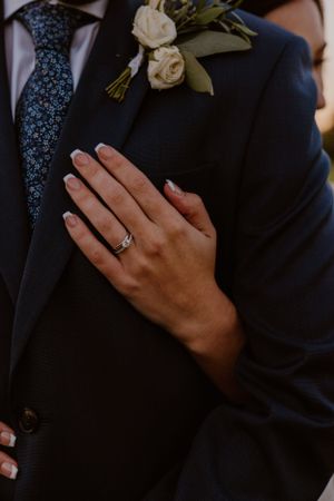 Close-up of engagement ring and white rose boutonniere on navy suit jacket.