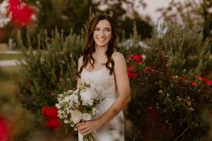 Bride in lace gown smiling with white bouquet among blooming red roses.