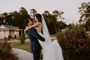 Bride in white dress and groom in navy suit embrace in a garden with red flowers.