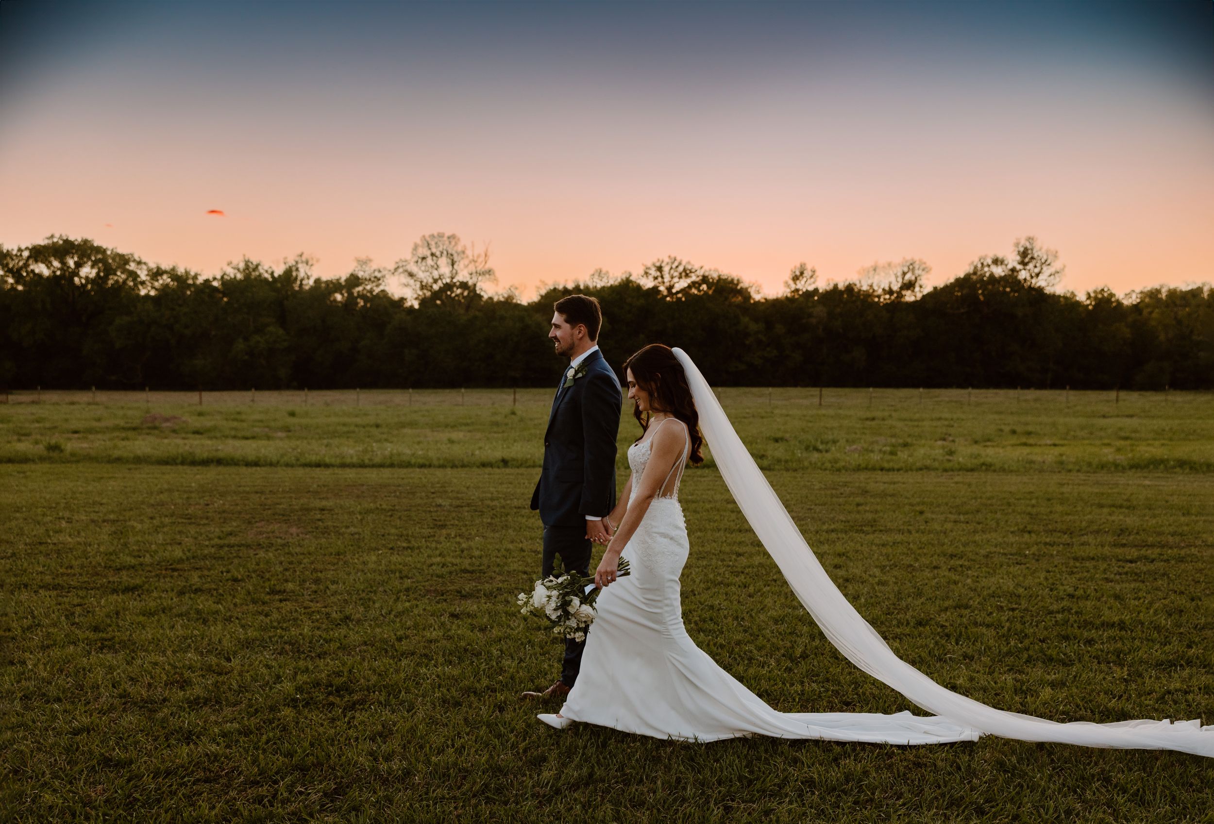 Newlyweds with long trailing veil walk together in open field at sunset.