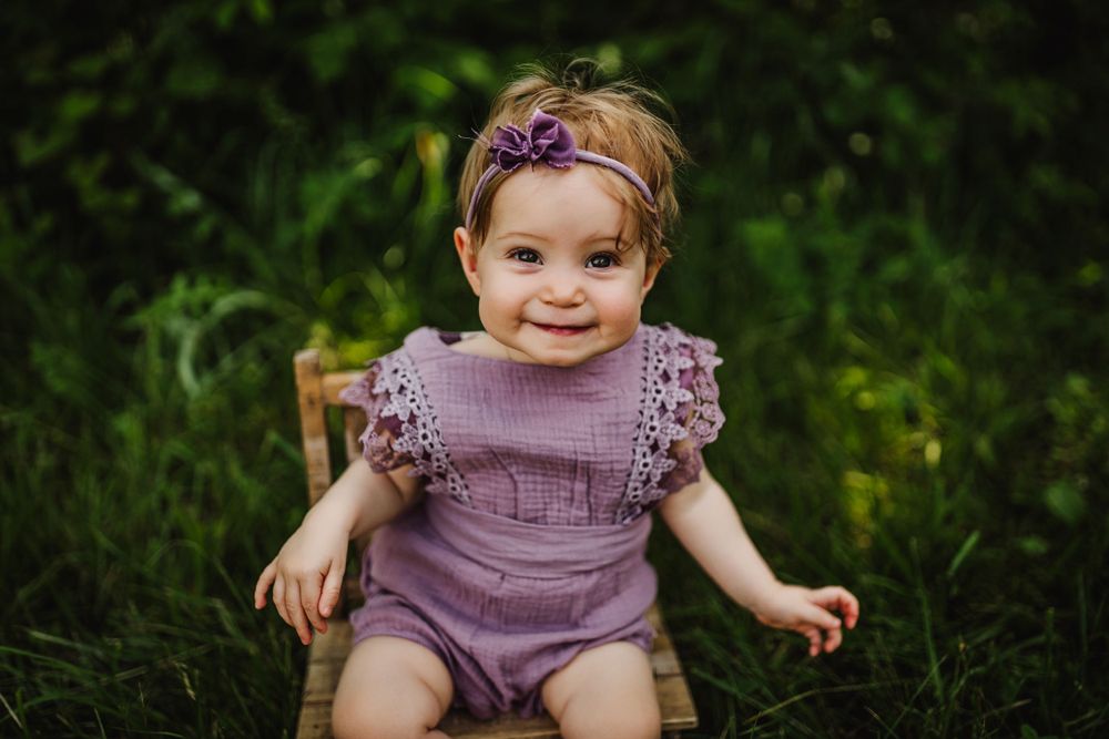 Baby in purple ruffled romper sits in wooden chair against green garden background.