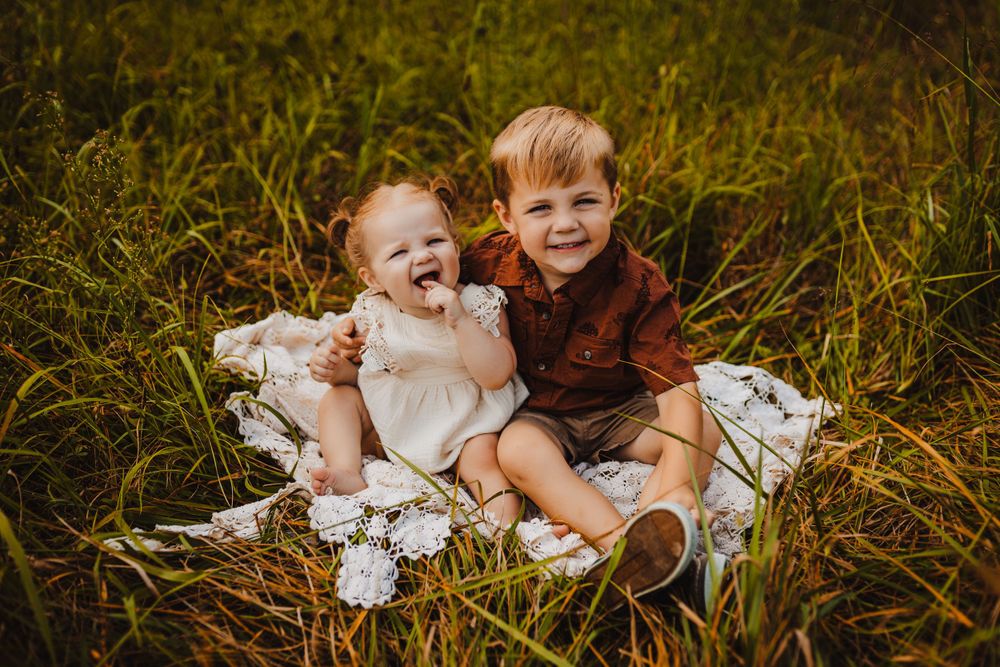 Two young children sitting together on blanket in tall grass field.