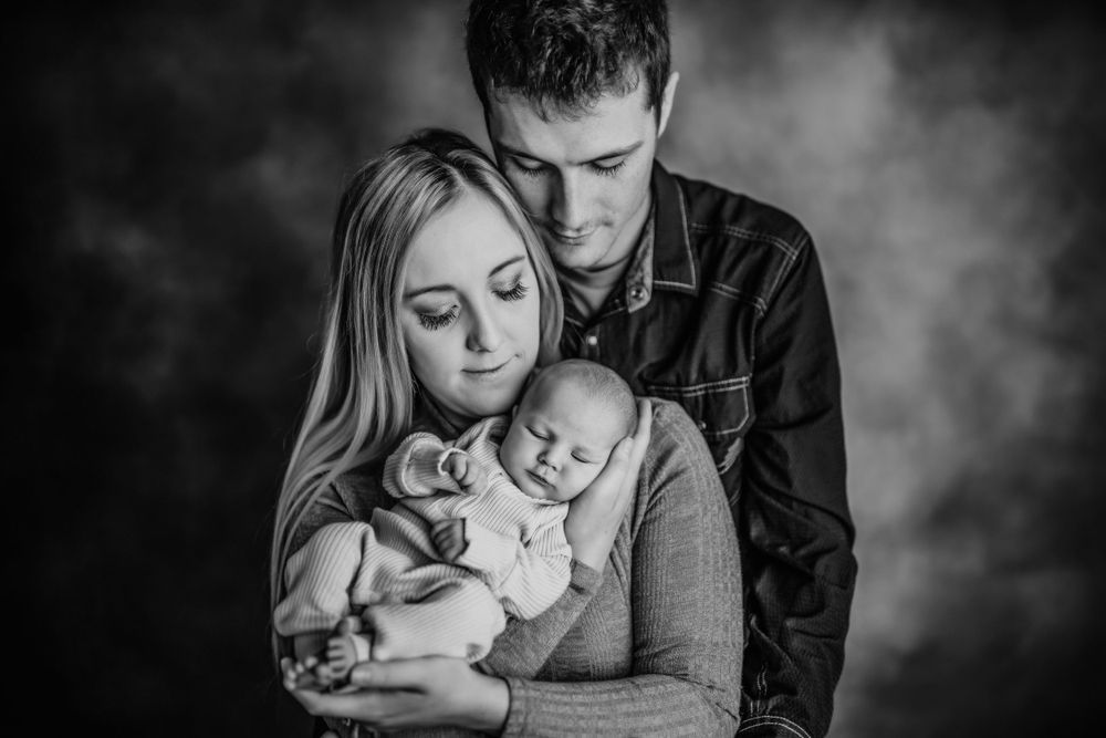 An intimate black and white portrait of parents with their newborn baby against a dark background.