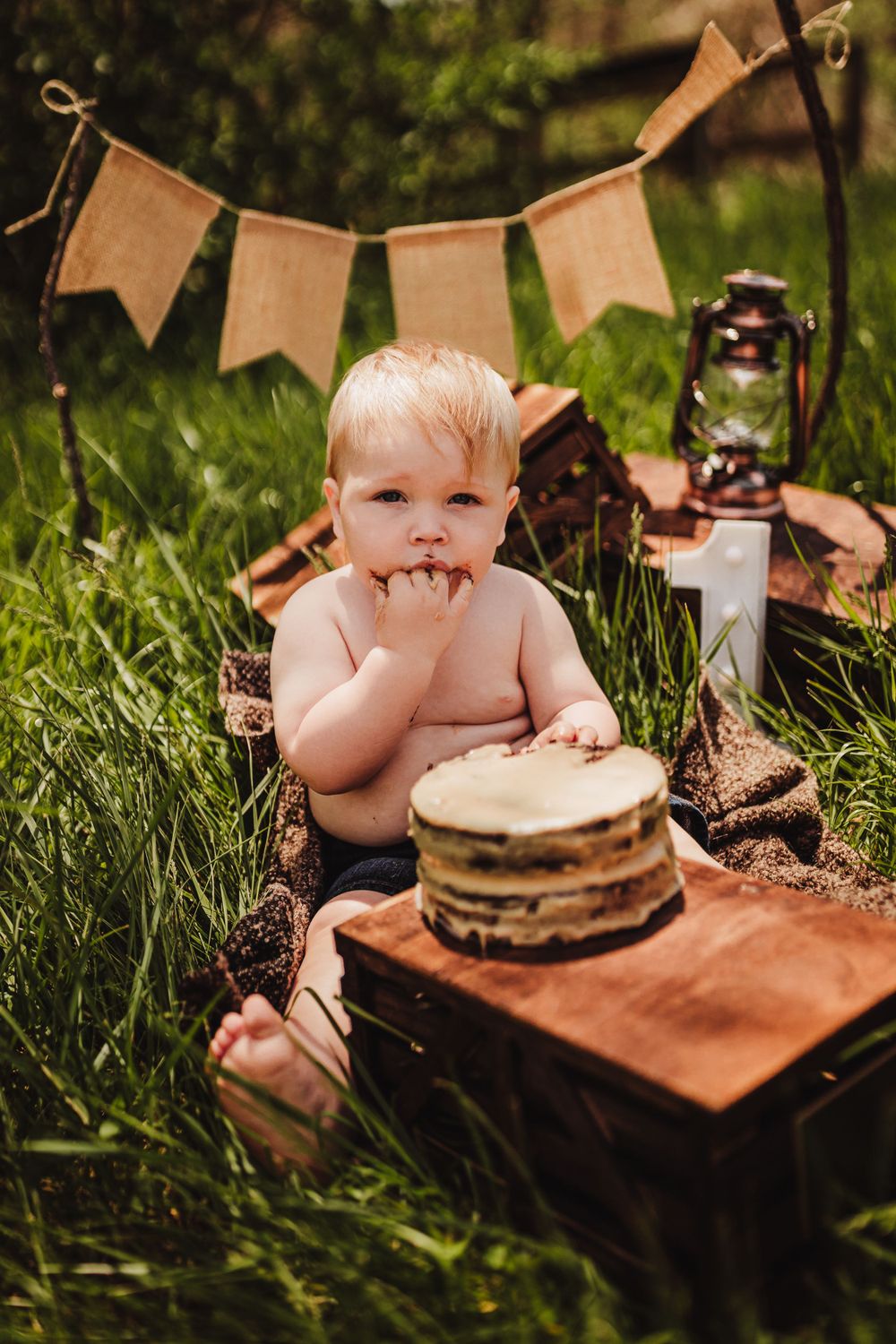 Outdoor cake smash photo setup with rustic decor including burlap banner and lantern in green grass.