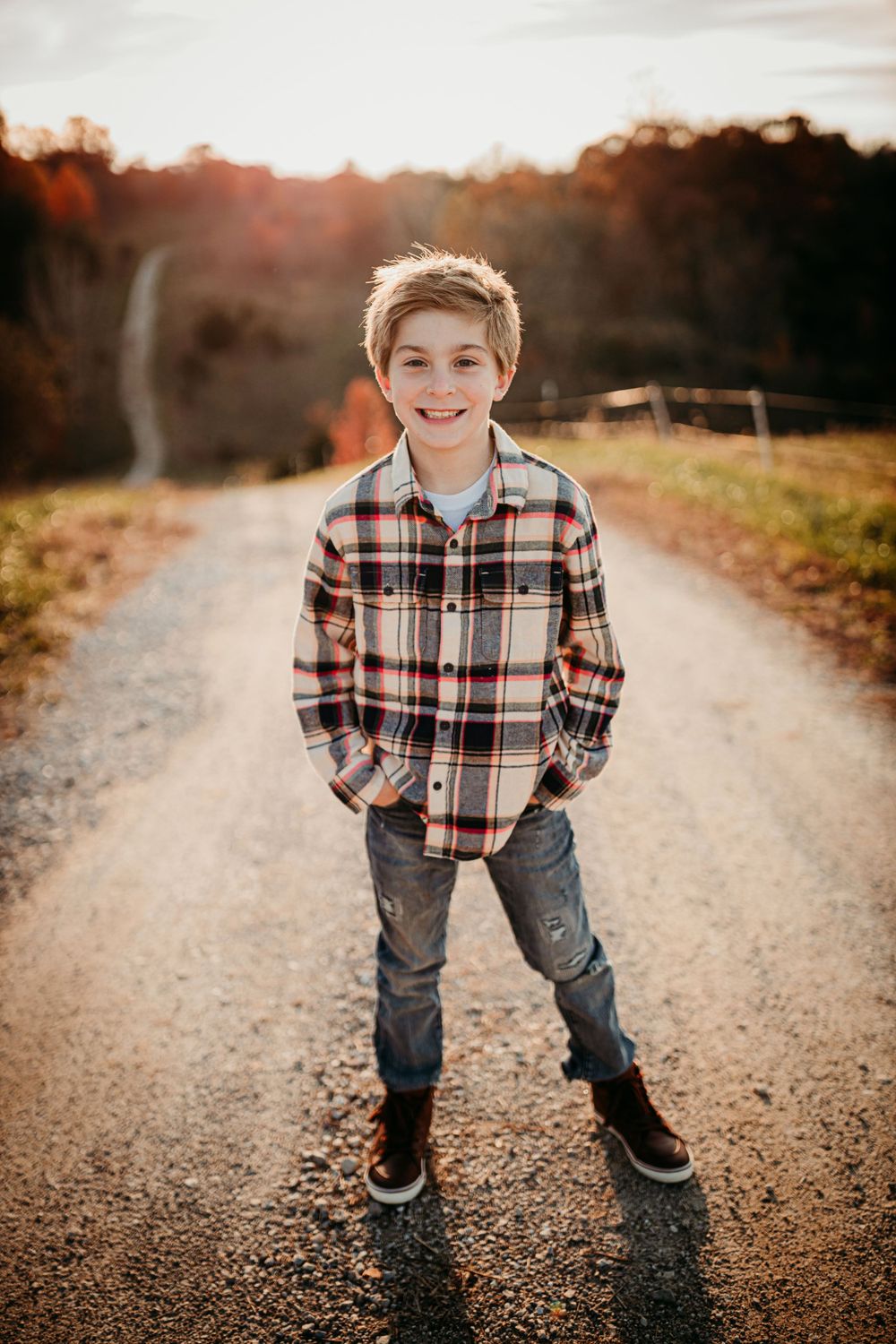 Young boy in plaid shirt smiling on dirt road at sunset.