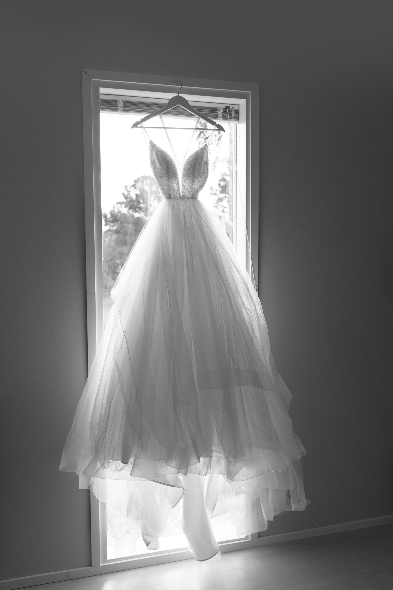 An elegant white wedding dress hanging in window backlit by natural light.