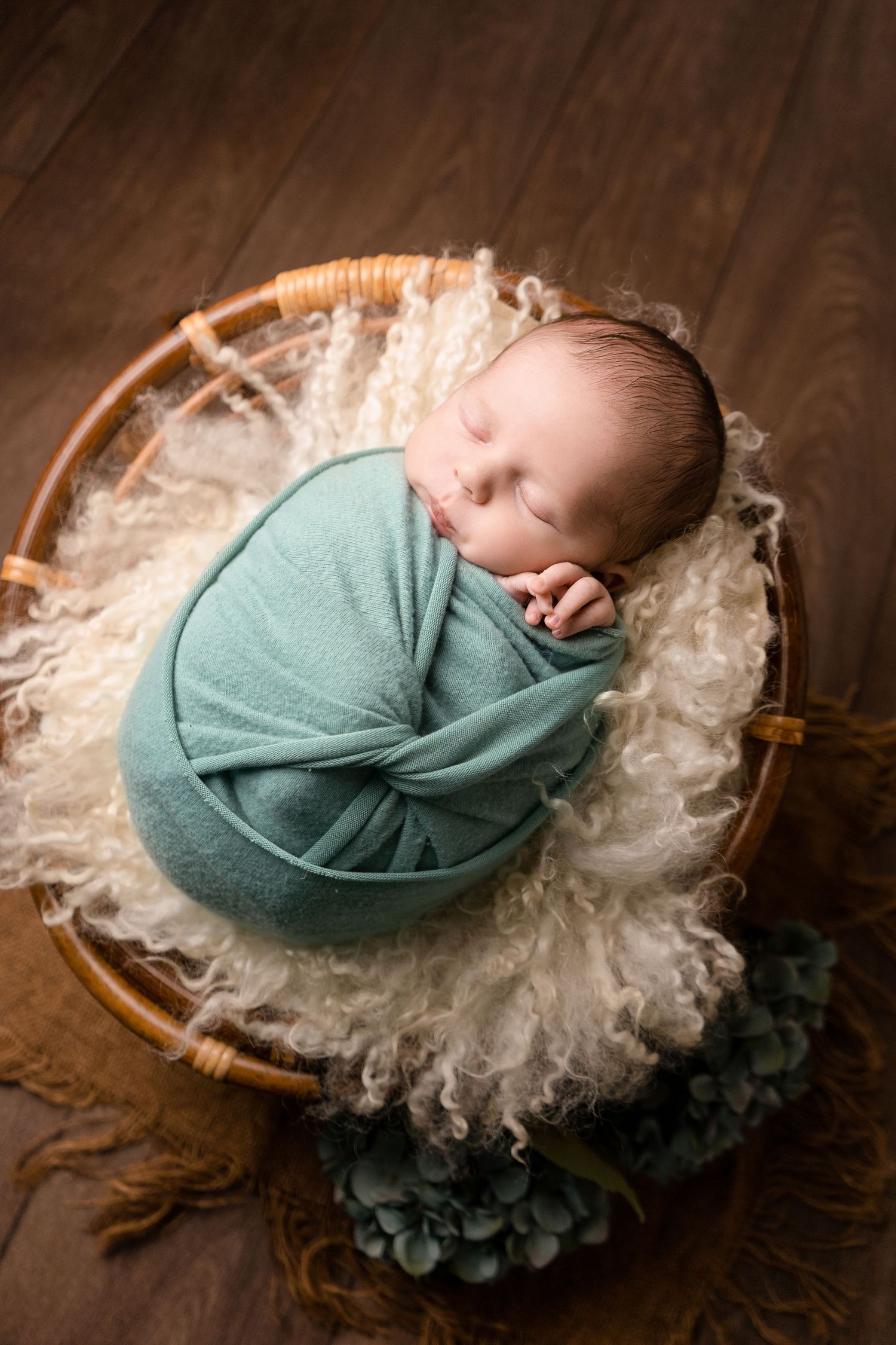 Baby boy sleeping wrapped in sage green on rattan chair, on brown wooden floor at newborn photoshoot in Doncaster.
