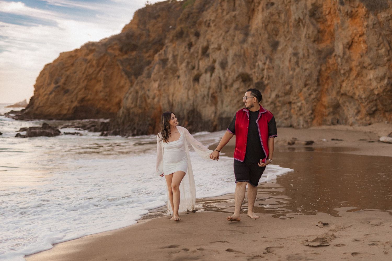 Couple walking hand in hand on sandy beach with dramatic cliffs in background at sunset.