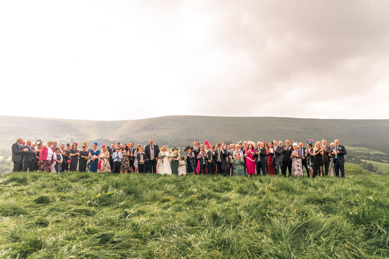 Large group photo of wedding guests lined up on a grassy hilltop with scenic countryside in background.