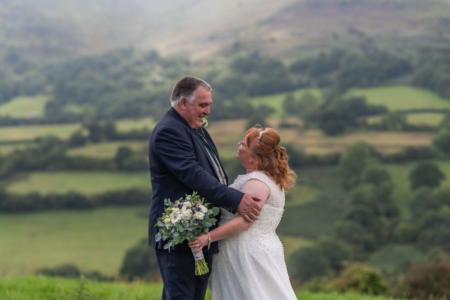 Couple embraces on hilltop with rolling countryside views in background.