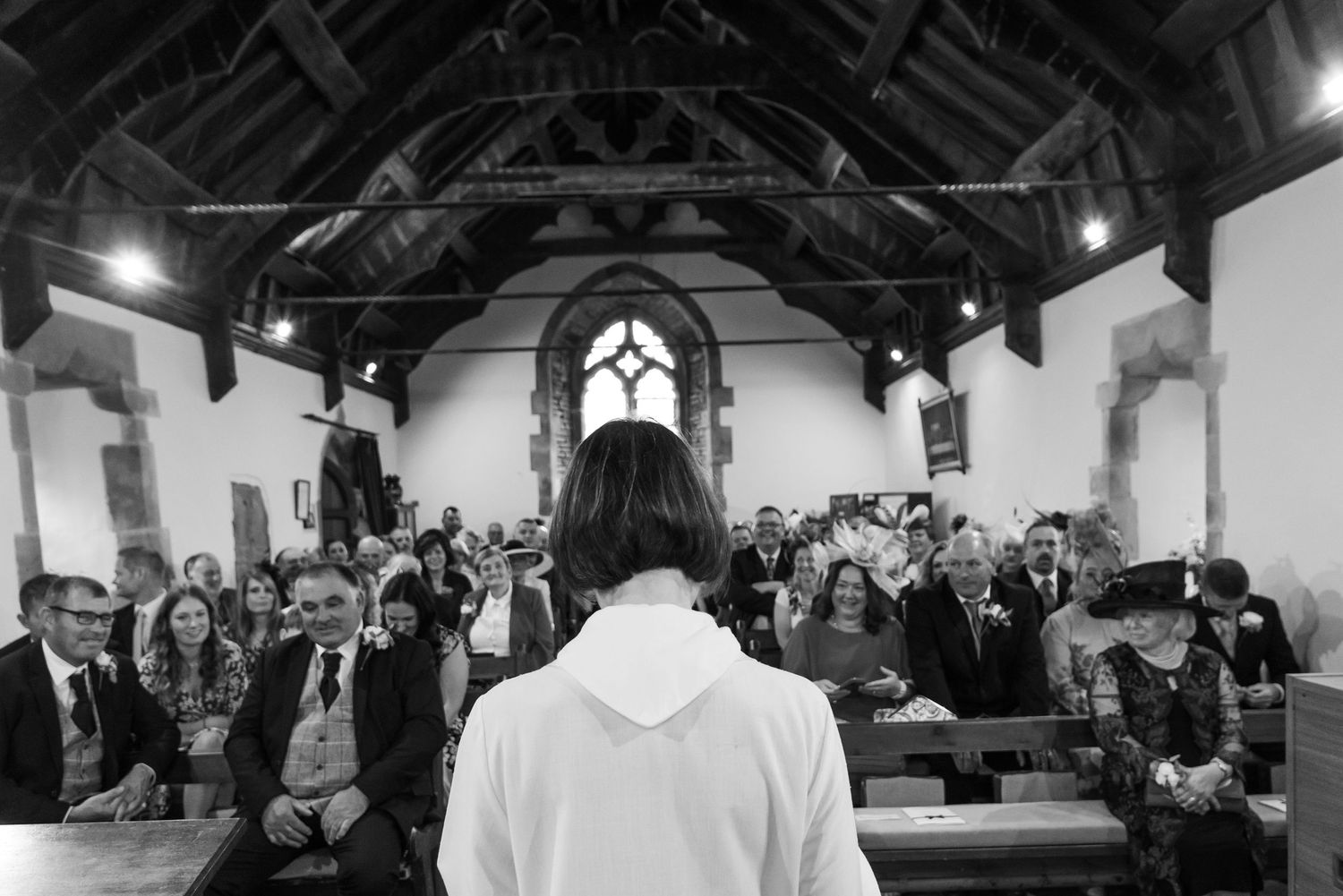 Black and white photo of person facing congregation inside historic church with wooden beams.