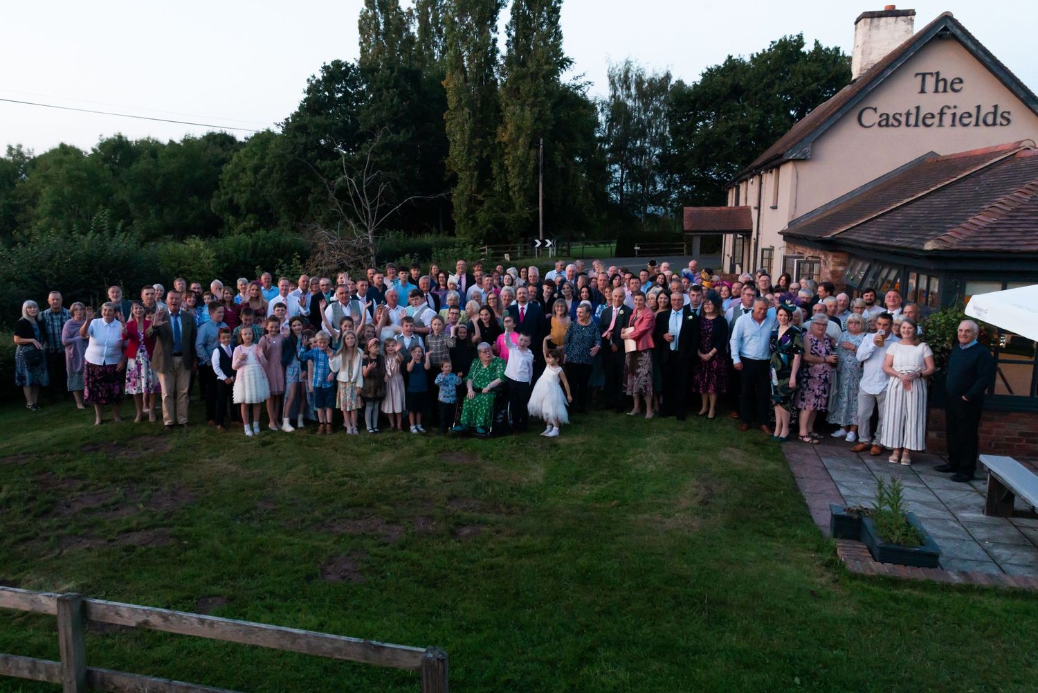 Large wedding party group photo outside The Castlefields venue at dusk.