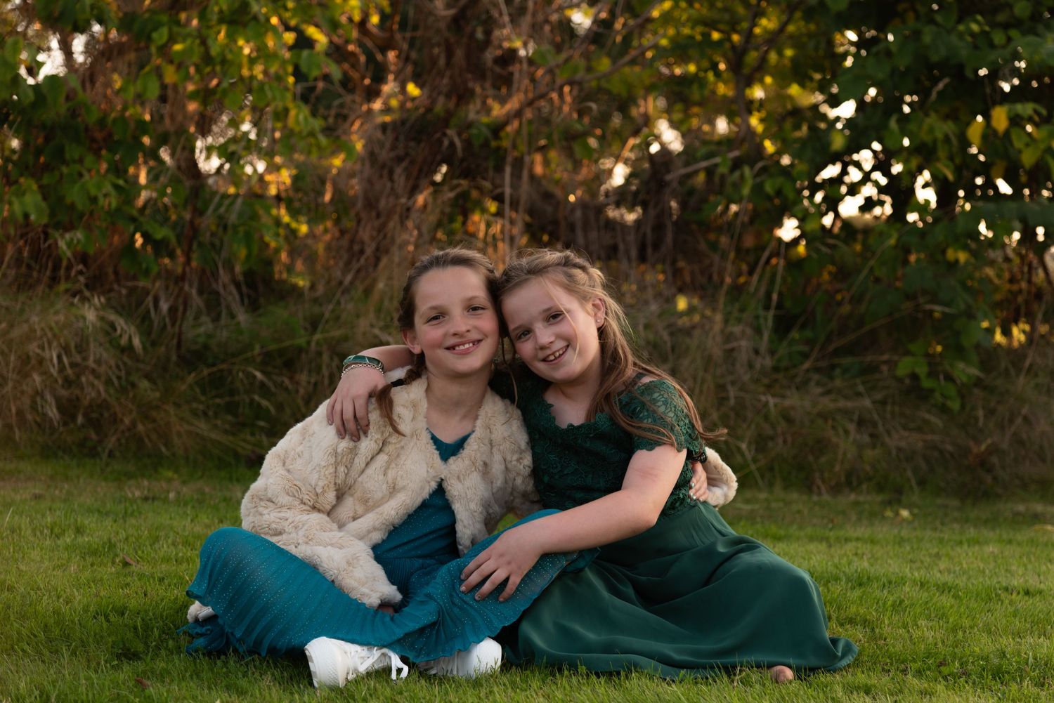 Two young friends wearing cozy fall outfits sit together smiling on green grass with autumn foliage in background.