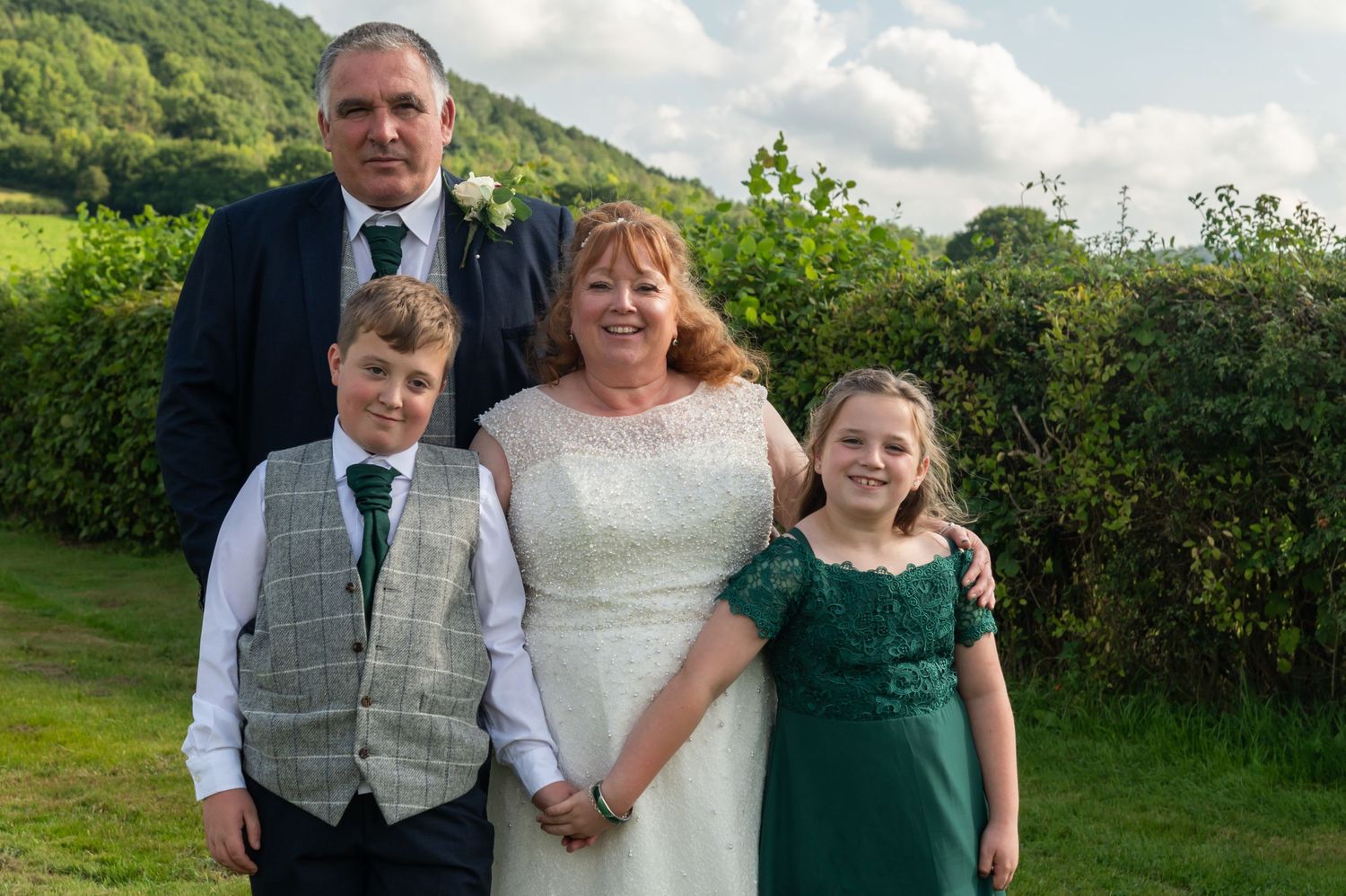 Wedding party poses together in front of green hedges and rolling hills.