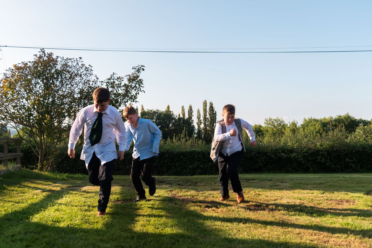 Wedding guests running playfully across a sunlit lawn during outdoor celebration.