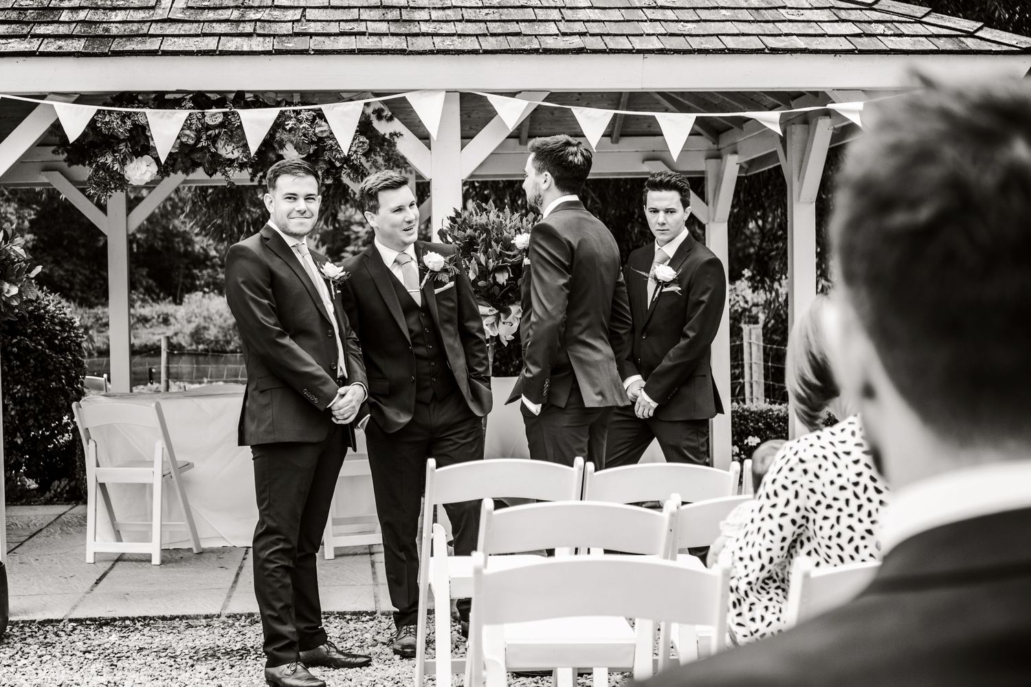 Black and white photo of wedding party gathered under a pavilion with white chairs.