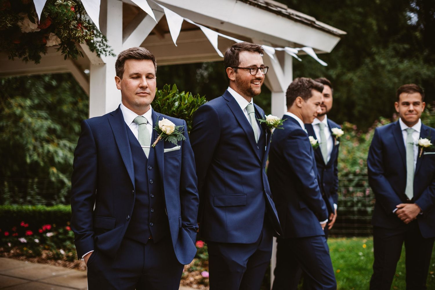 Wedding party in coordinated navy suits standing under a decorated pavilion.