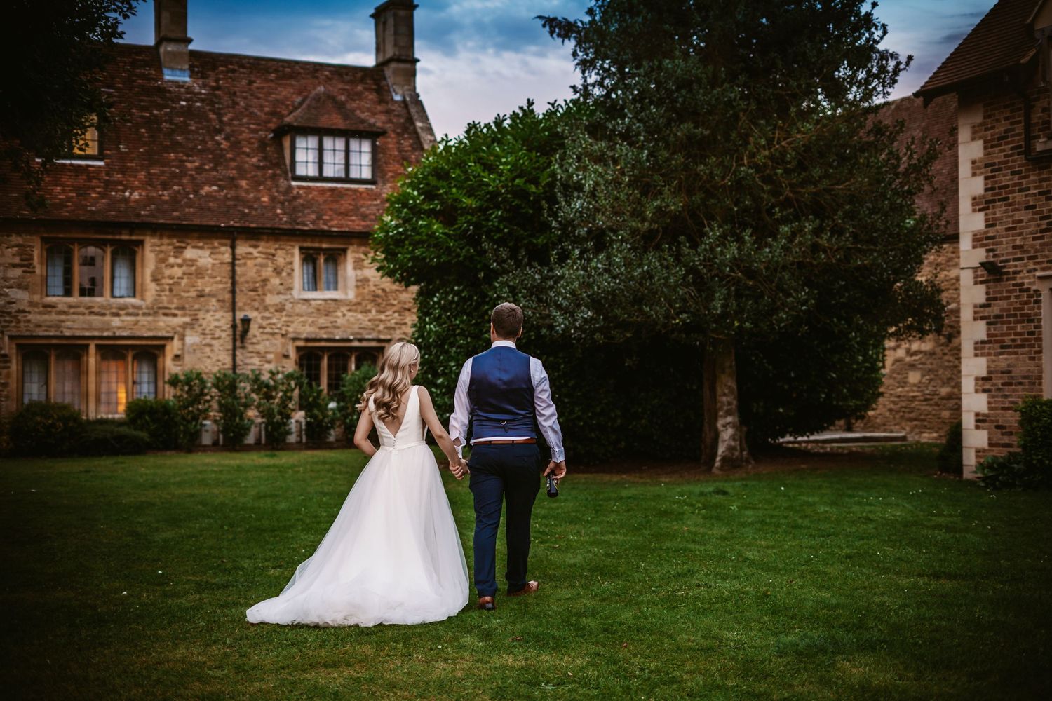 A romantic wedding couple walks across a lush green lawn toward a stone country house on their special day.