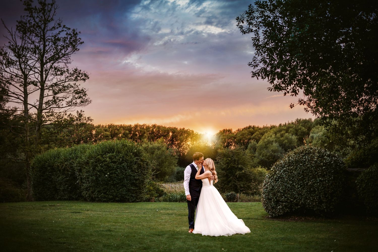 A wedding couple stands together against a stunning sunset backdrop surrounded by manicured garden hedges.