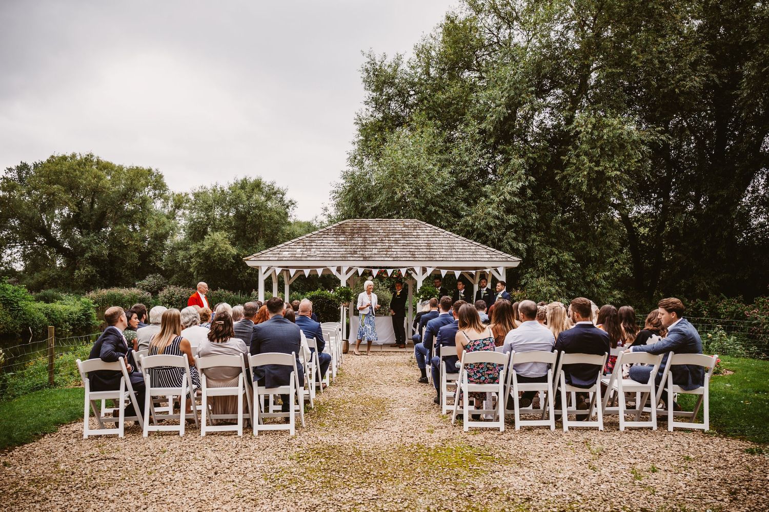 Outdoor wedding ceremony setup with white chairs and gazebo on a gravel path surrounded by trees.