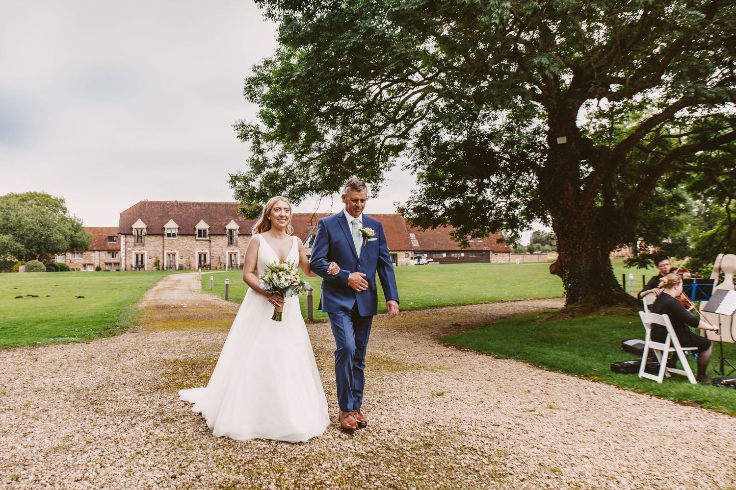 Newlyweds walking down gravel path with historic building and large tree in background.