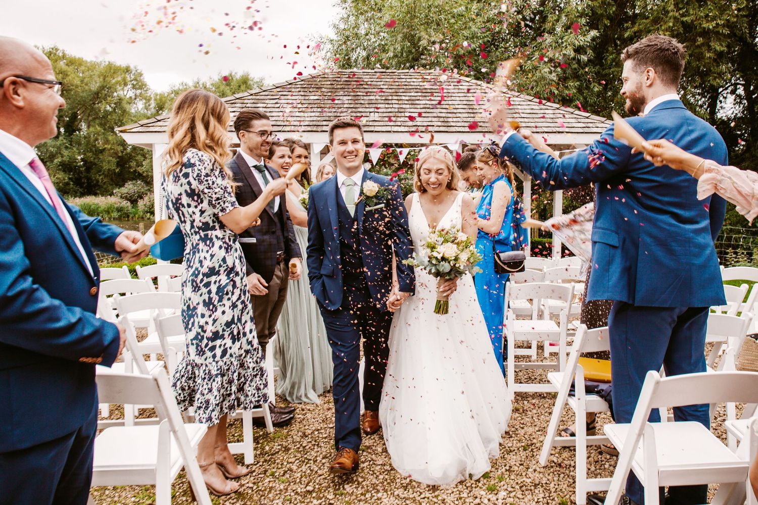 Wedding celebration with guests tossing confetti at couple walking down garden aisle.