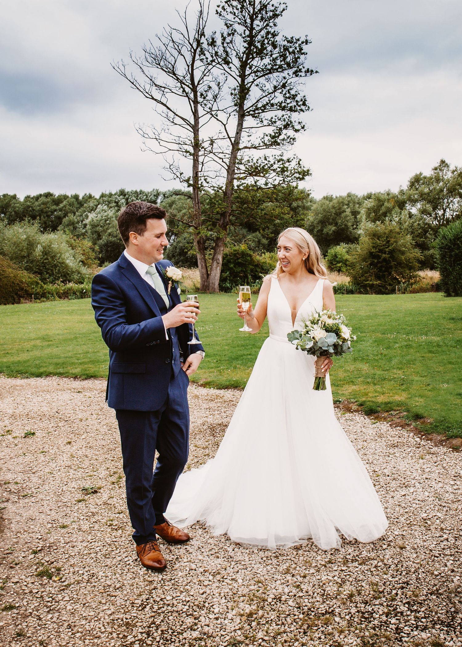 Wedding couple sharing champagne toast on gravel path in rustic outdoor setting.