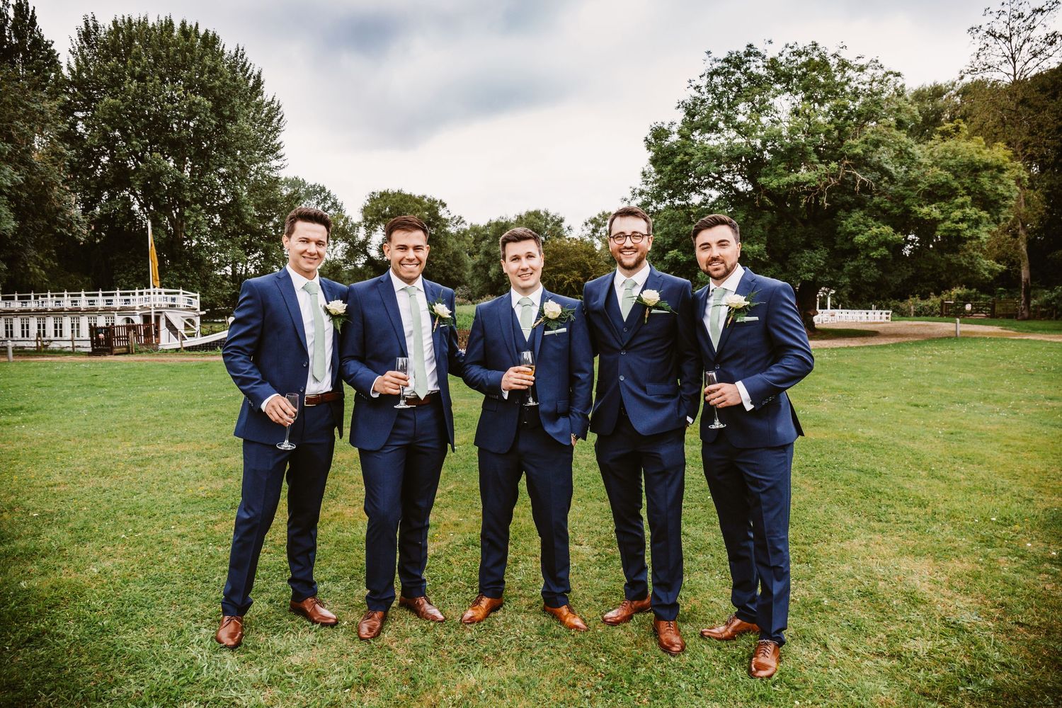 Groomsmen in navy suits and brown shoes pose on garden lawn.