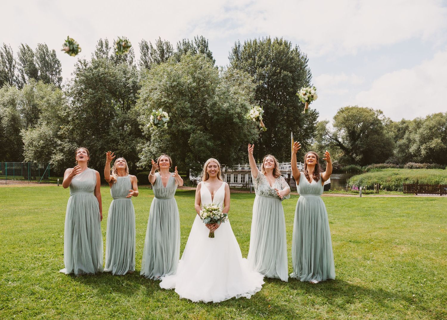 A wedding party in light blue bridesmaid dresses poses together on a grassy lawn with trees in the background.