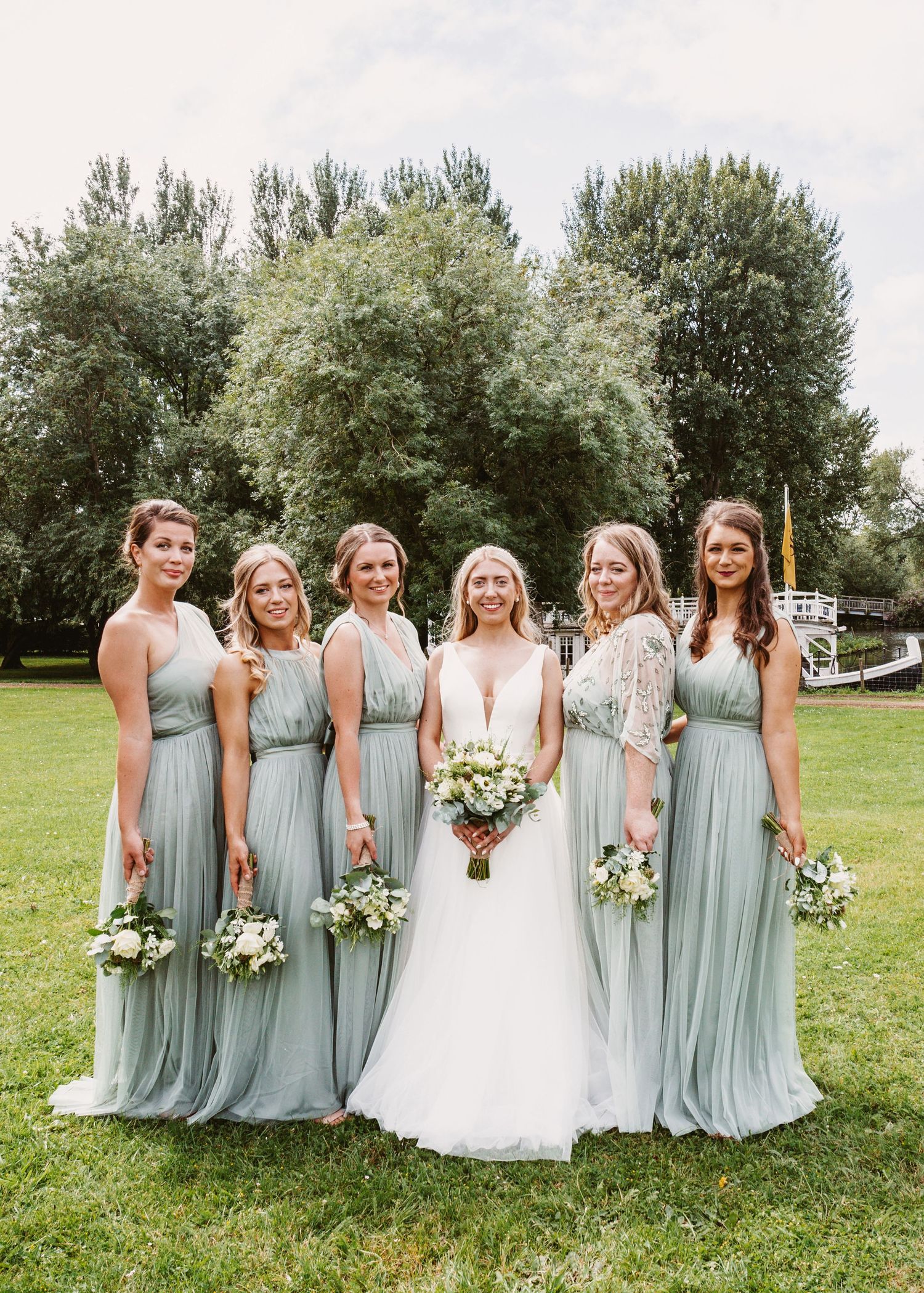 Bridal party in sage green dresses poses with bouquets on garden lawn.