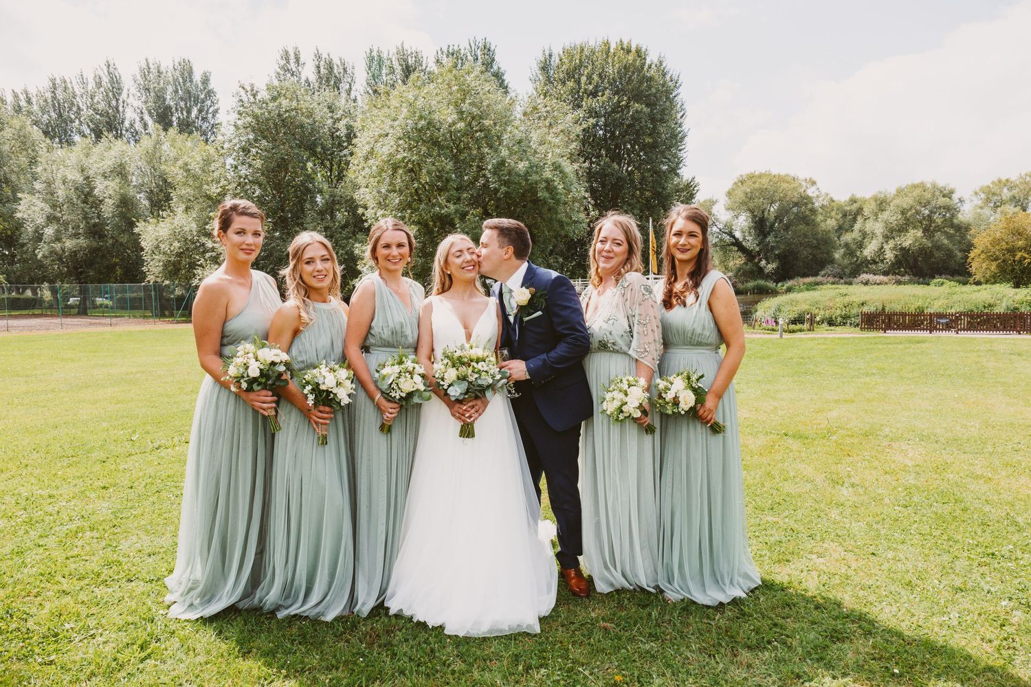 Wedding party gathers for a formal photo on green lawn with bridesmaids in elegant sage green gowns.