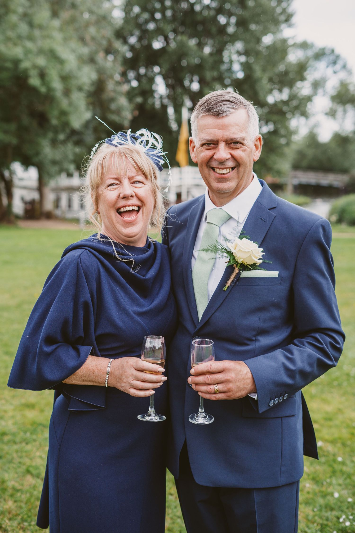 A couple in formal attire shares a laugh while holding champagne glasses at an outdoor event.