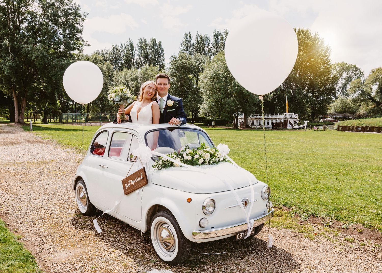 Wedding couple sits in decorated classic white Fiat with large white balloons in countryside setting.