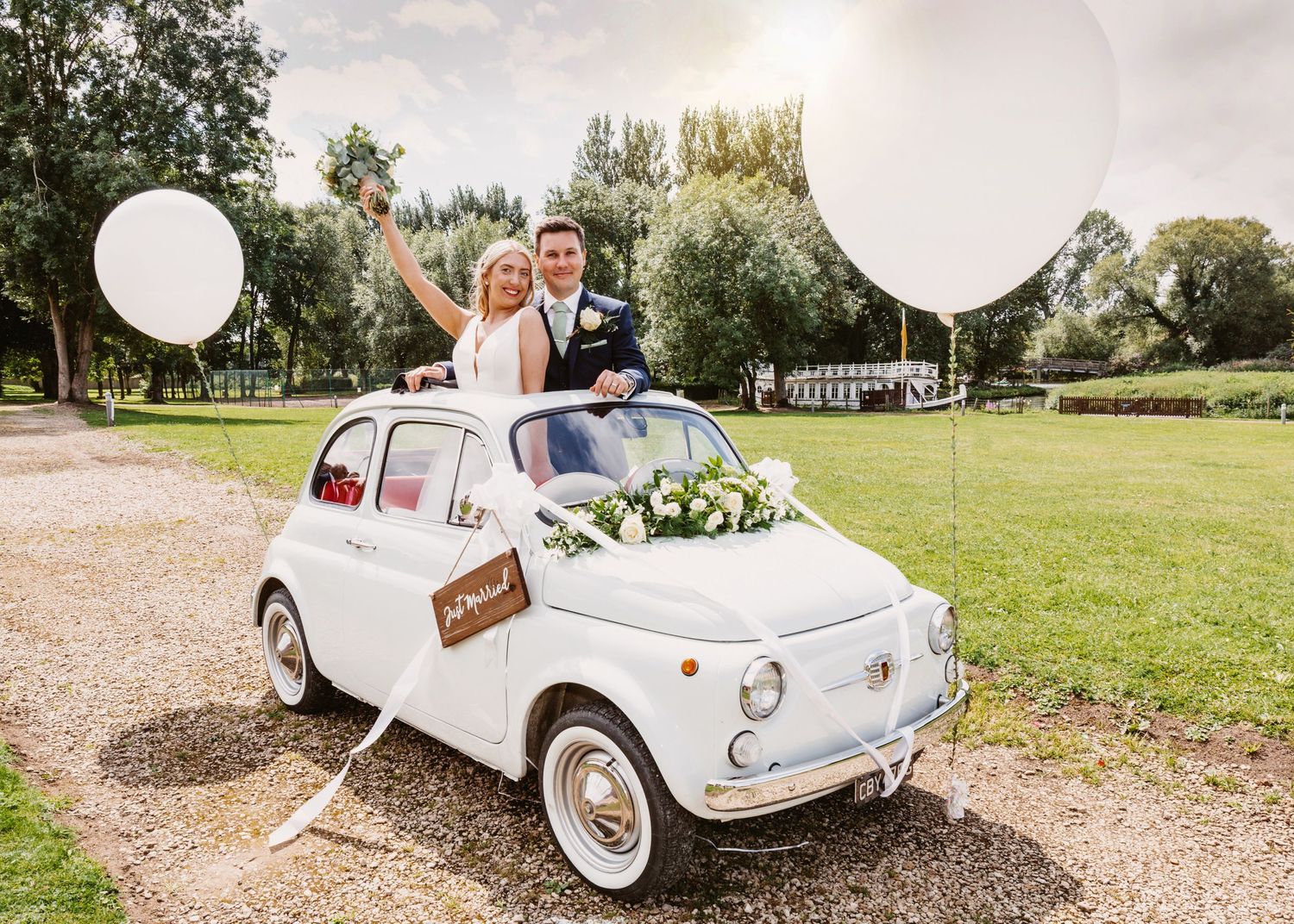 Newlyweds pose in vintage white Fiat 500 decorated with balloons on a country path.