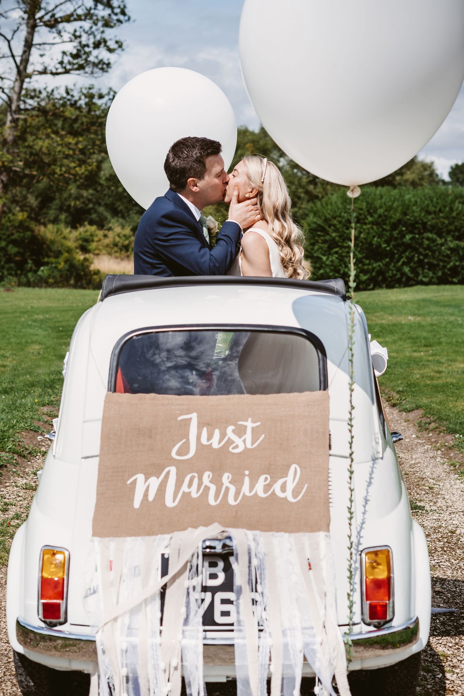 Wedding couple shares romantic moment in vintage Fiat decorated with Just Married sign and balloons.