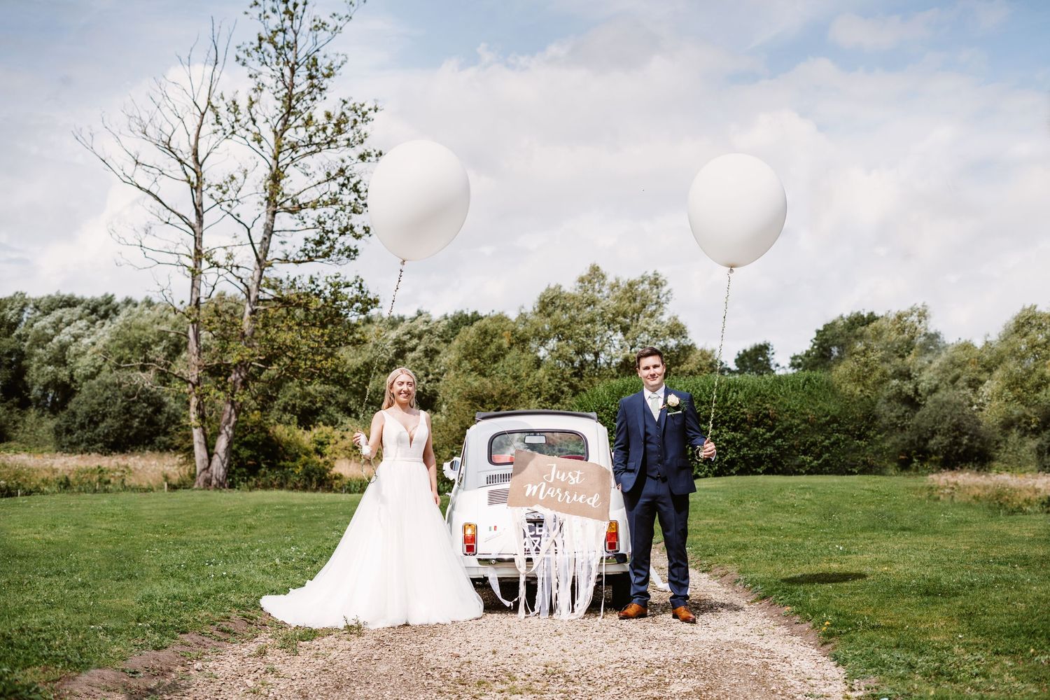 Wedding couple poses beside decorated vintage white Fiat with balloons in countryside setting.