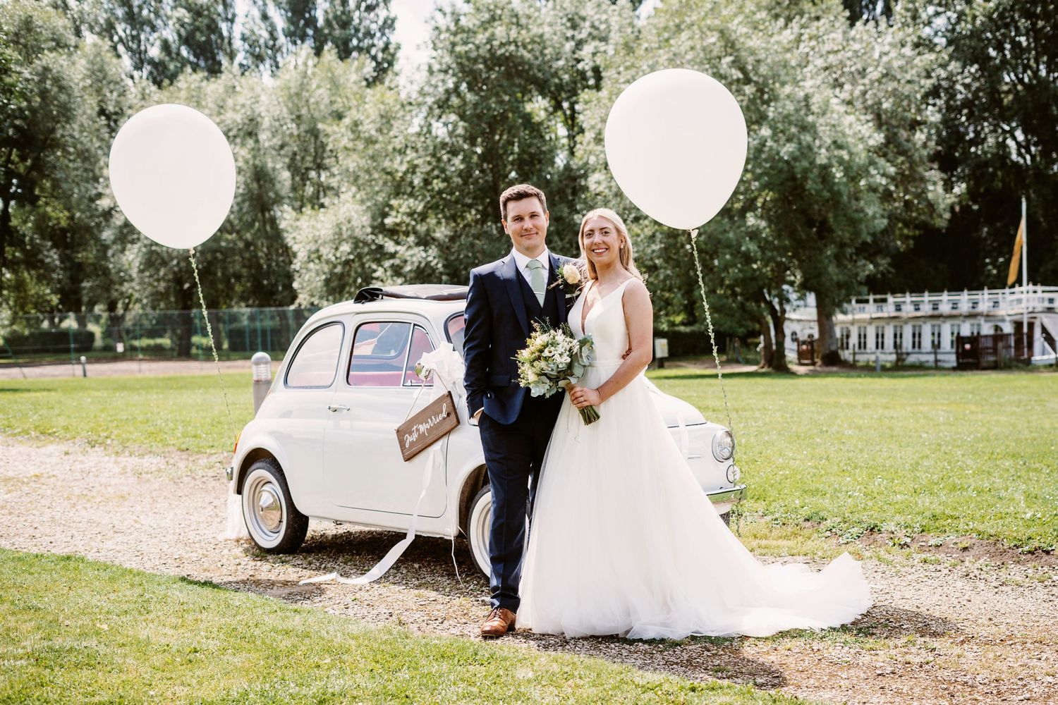 Wedding couple poses with white balloons next to vintage Fiat 500 car on a sunny day.