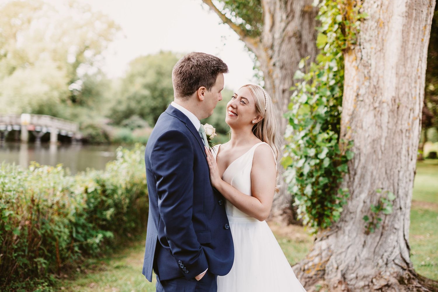 Romantic wedding couple shares intimate moment near ivy-covered tree by water.