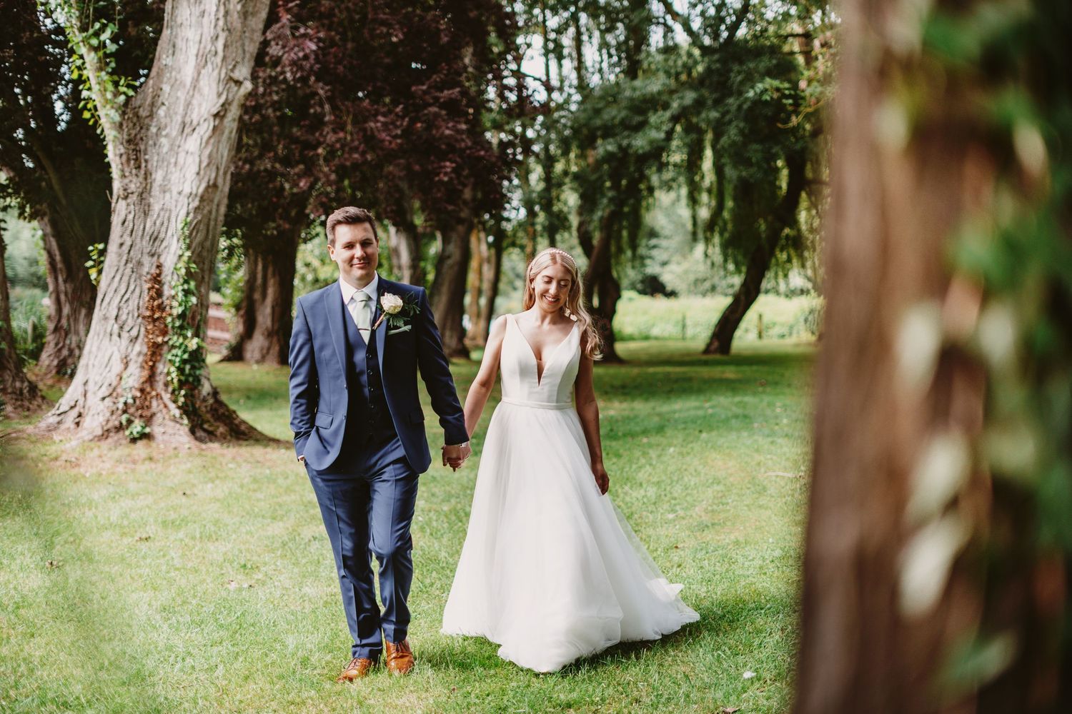 Couple walks hand in hand through wooded park in wedding attire.