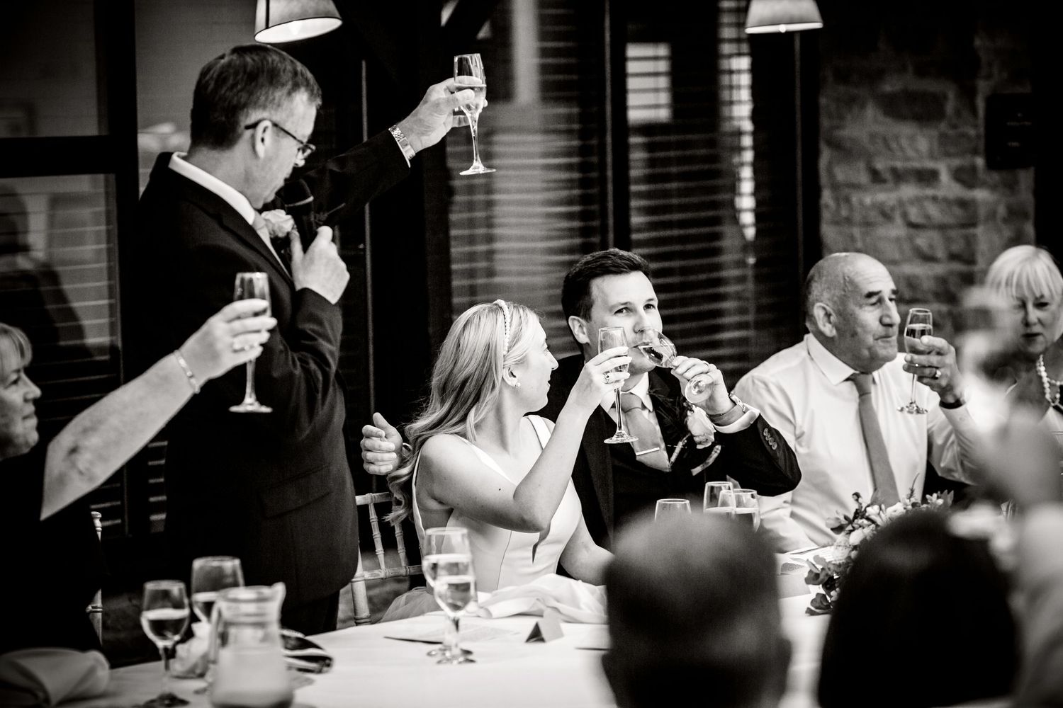 Wedding guests raise glasses in celebration during black and white reception moment.