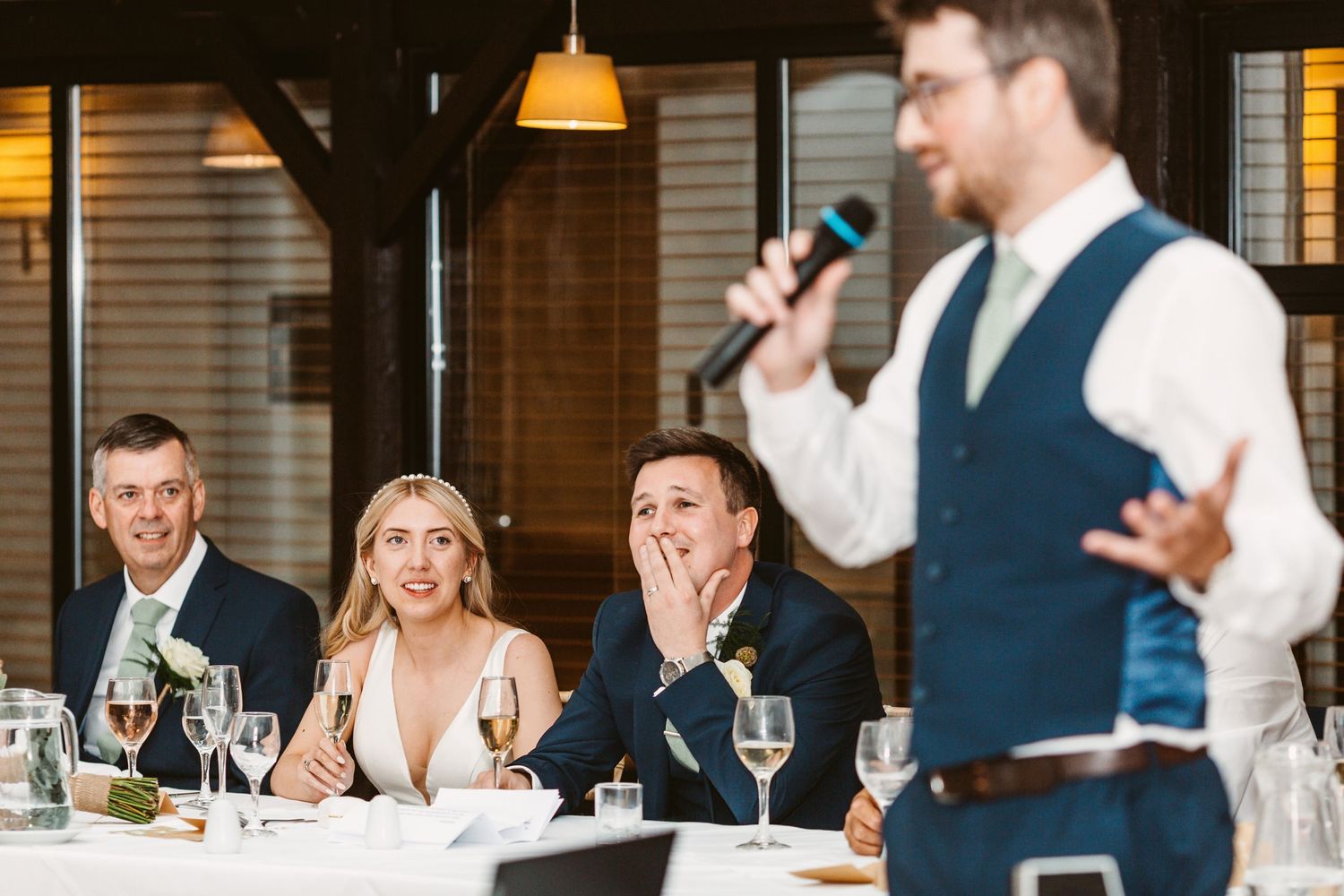 Wedding reception guests laugh during speech while sitting at decorated dinner table.