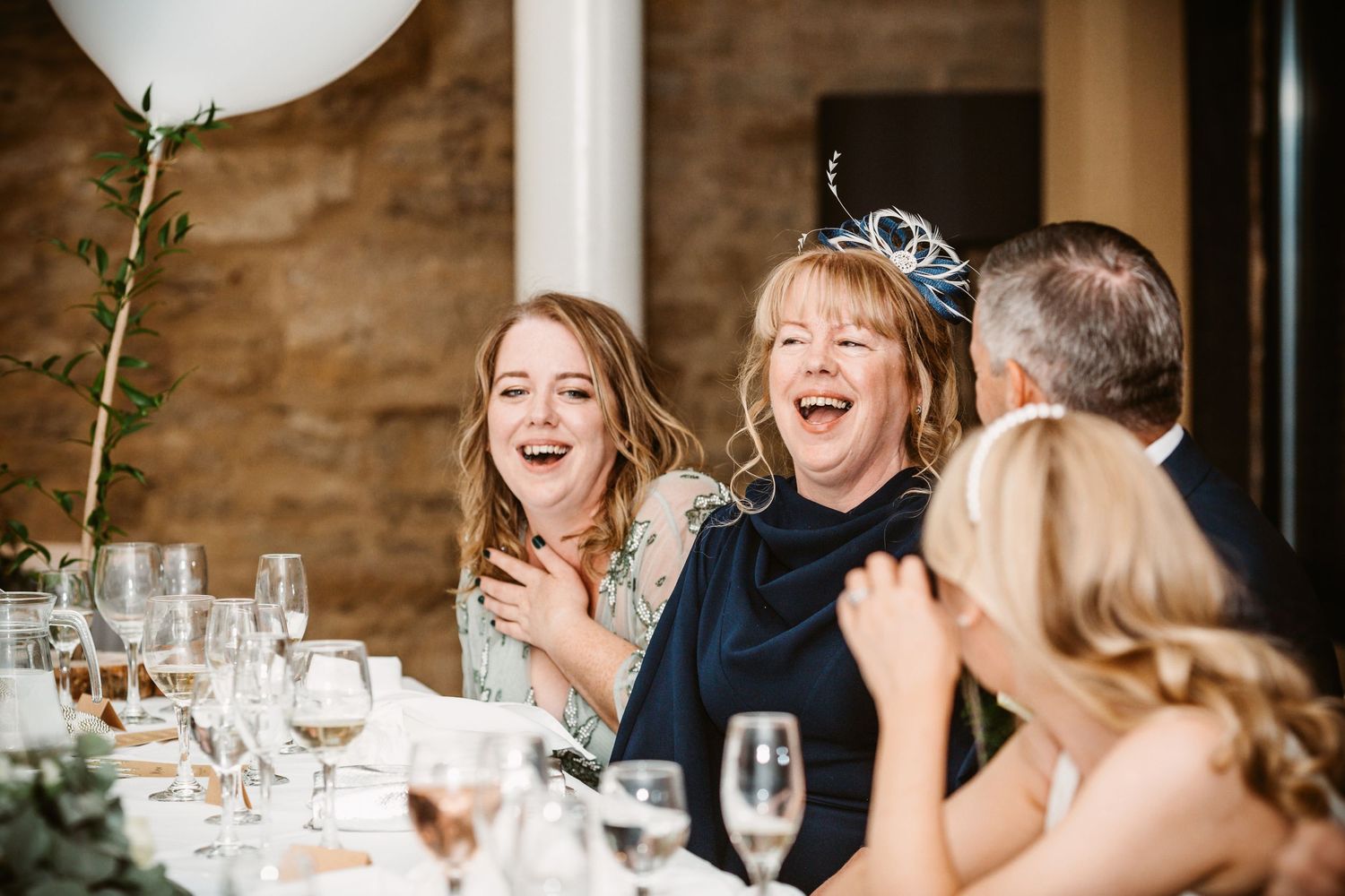 Joyful guests share laughter around an elegant dinner table with white balloons at a wedding reception.