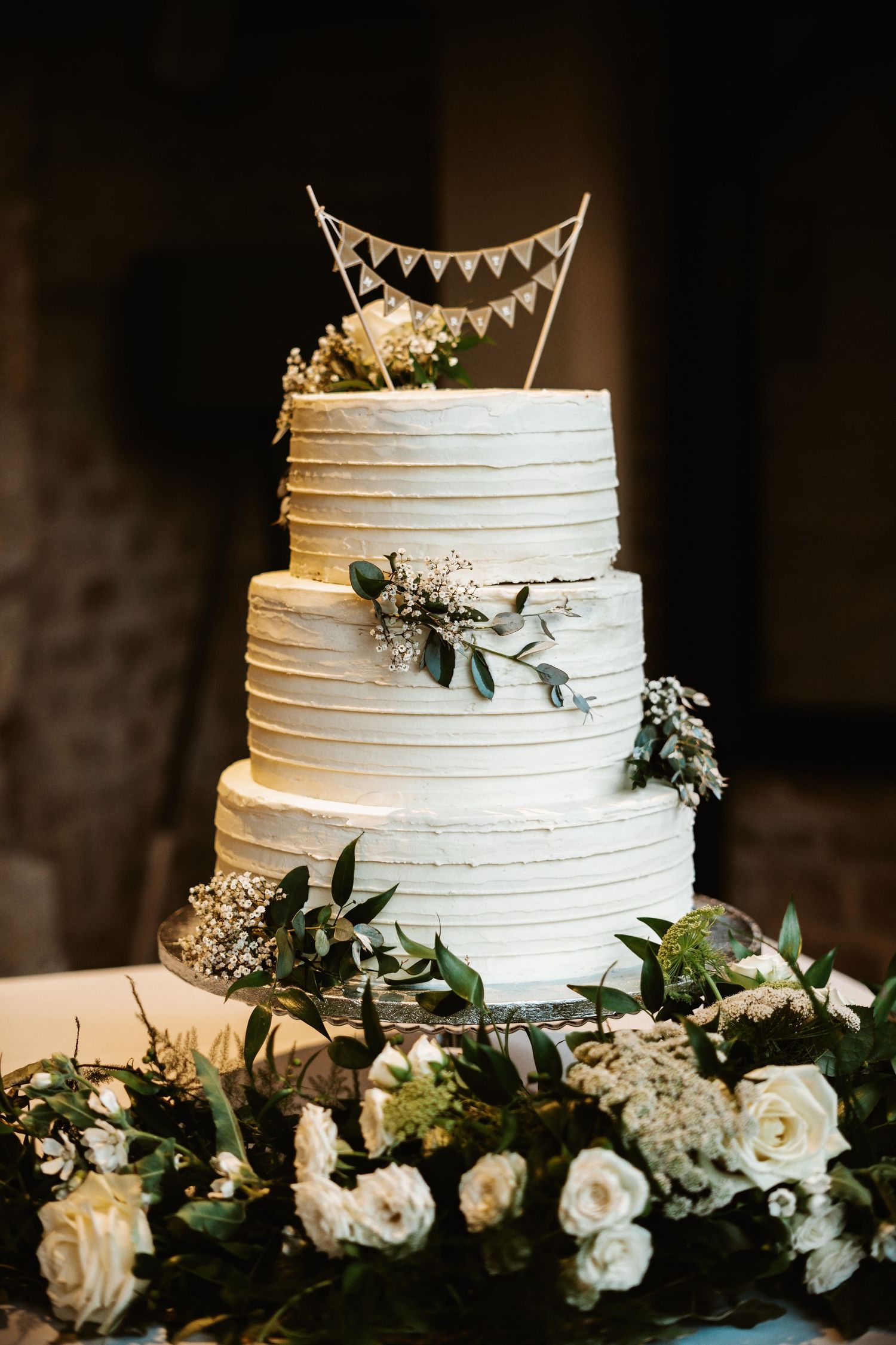 Three-tiered white wedding cake decorated with eucalyptus, roses and bunting topper on dark background.