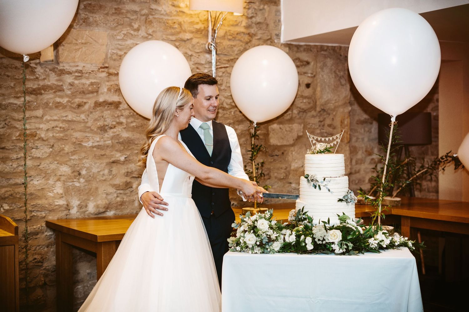 Couple cuts their elegant white wedding cake surrounded by balloons in rustic venue.
