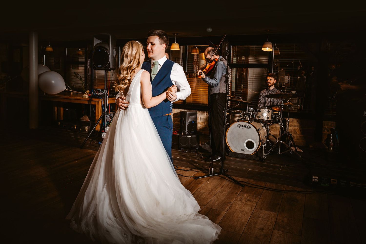 Couple shares first dance in rustic venue with band setup and wooden floors.