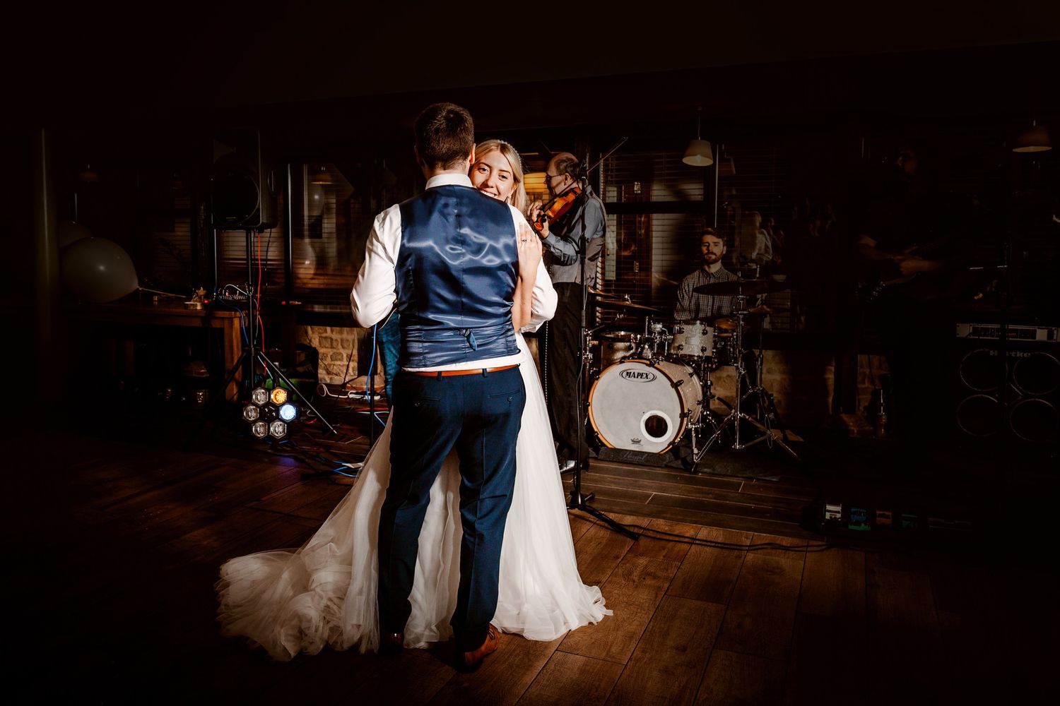 Wedding couple dances in intimate lighting with band equipment silhouetted in background.