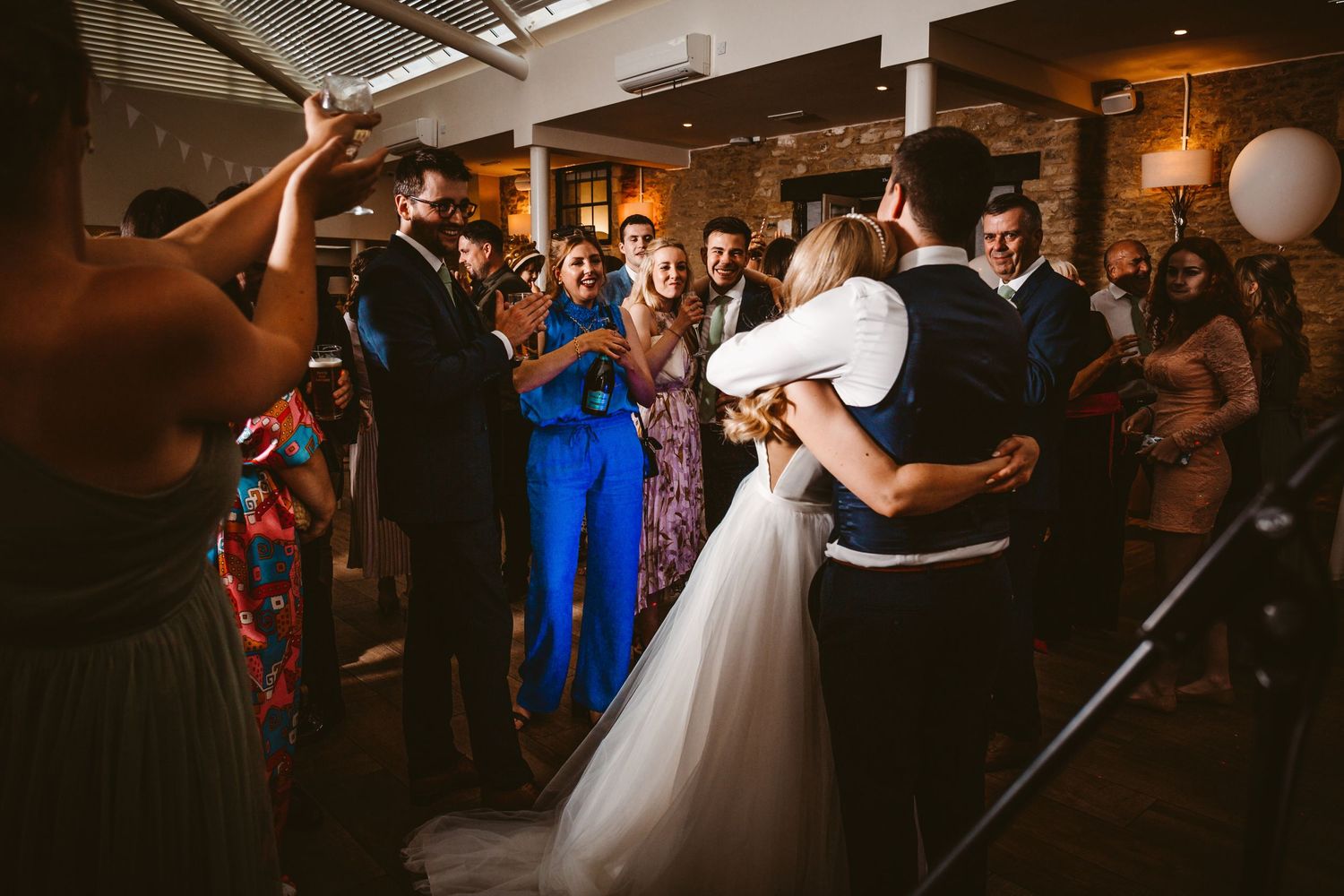 Wedding guests surround and cheer for the dancing newlyweds in a rustic indoor reception venue.