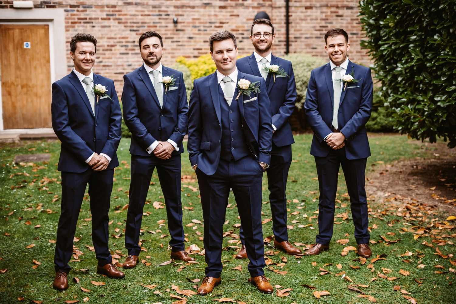 Groomsmen in matching navy suits and brown shoes pose together on autumn grass near a brick wall.