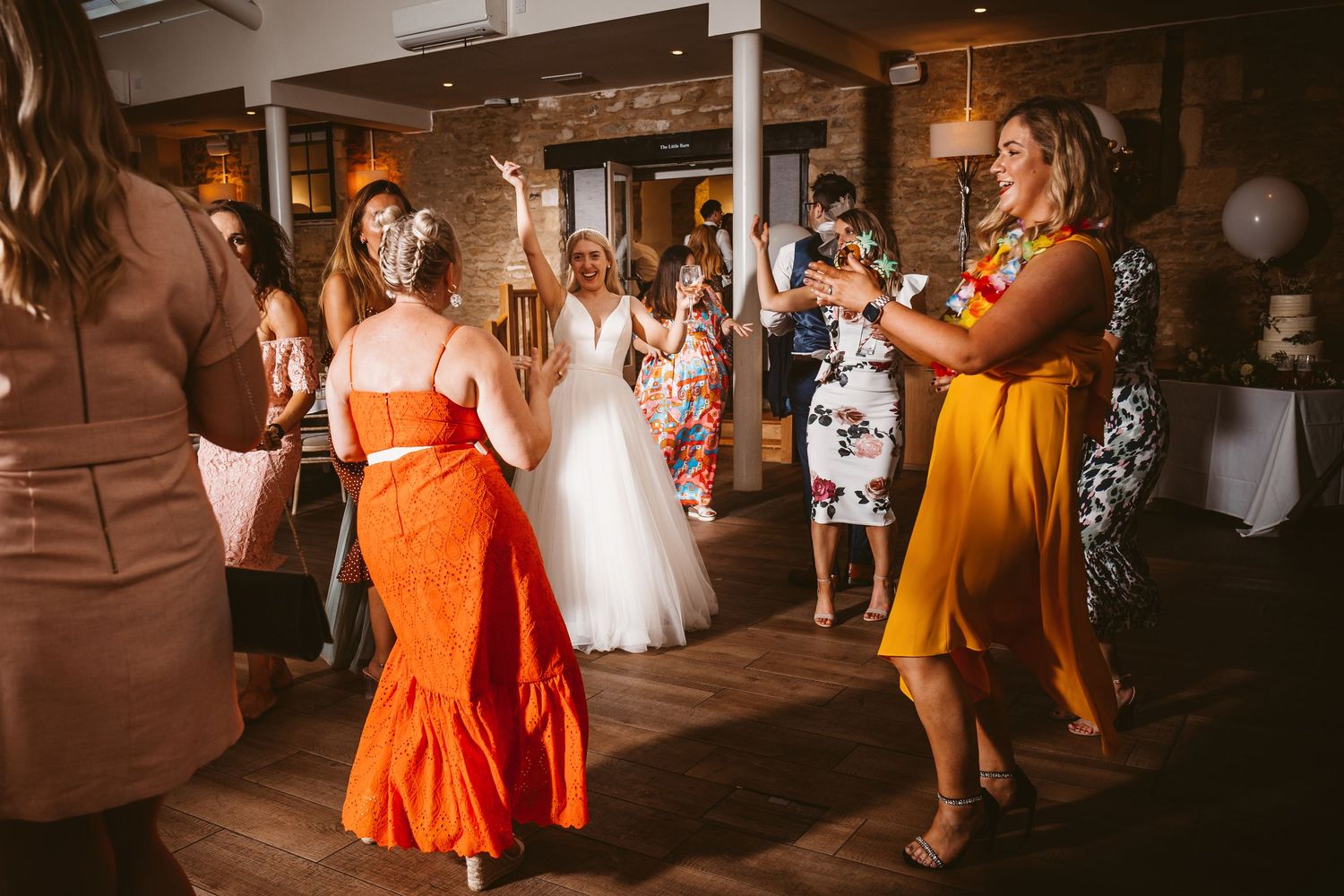 Wedding guests in colorful dresses dancing and celebrating at an indoor reception venue.