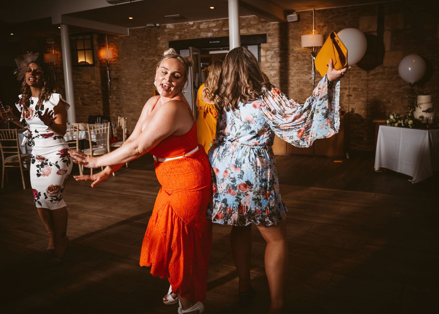 Guests in vibrant dresses dancing joyfully at a wedding reception with exposed brick walls.
